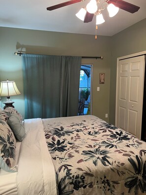 Une chambre à coucher comprenant un lit, un ventilateur de plafond, une table de chevet, une lampe, une porte et une fenêtre avec des rideaux.
