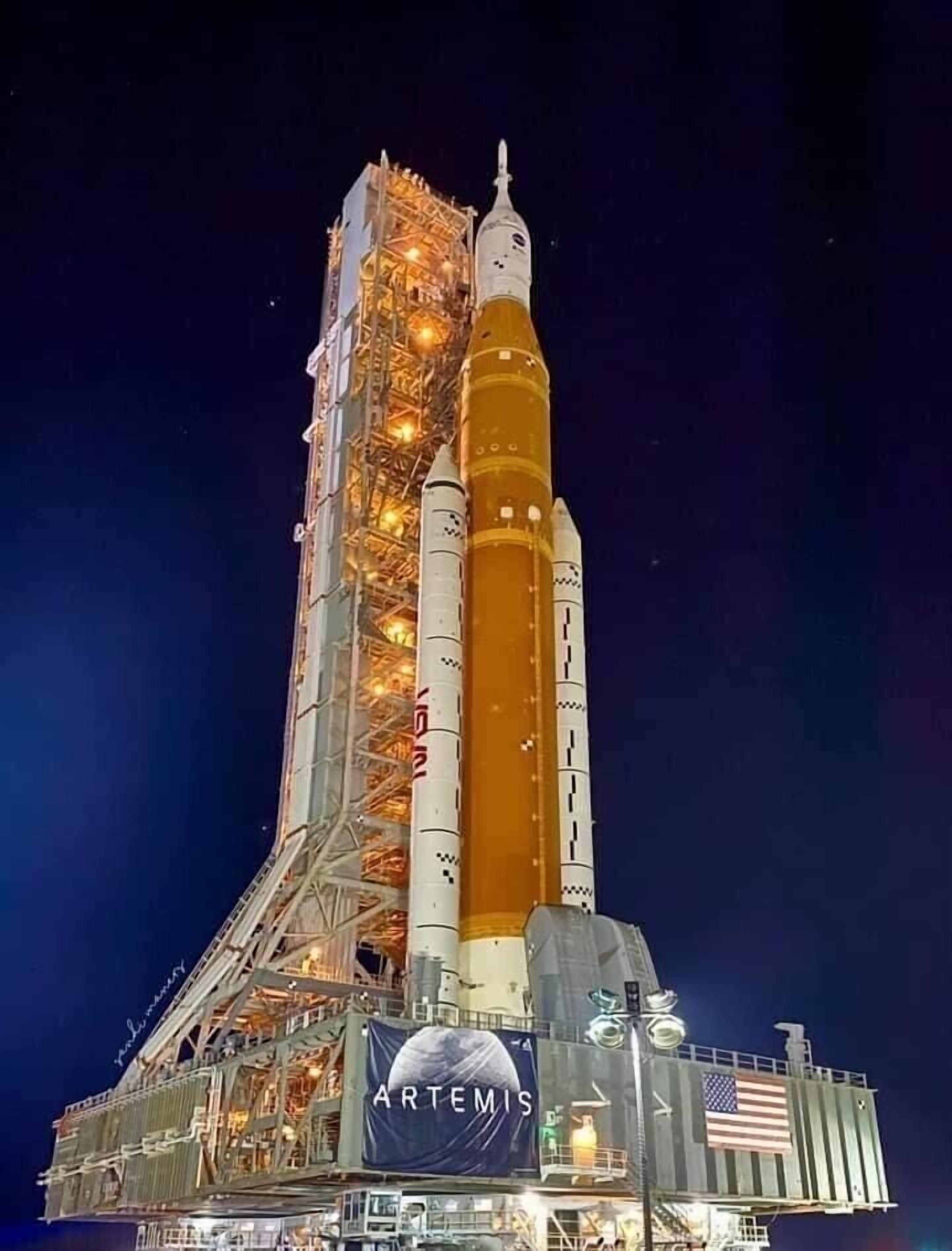 A tall rocket with a vertical orientation, attached to a launch structure with a visible American flag.
