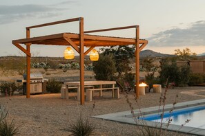 Una pérgola de madera con luces colgantes, una parrilla para barbacoa, una piscina y un paisaje desértico al fondo.