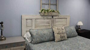 Ein Schlafzimmer mit einem Bett, einem Nachttisch, einer Lampe, einem Fenster mit Vorhang und einem Wandregal mit Blumen.