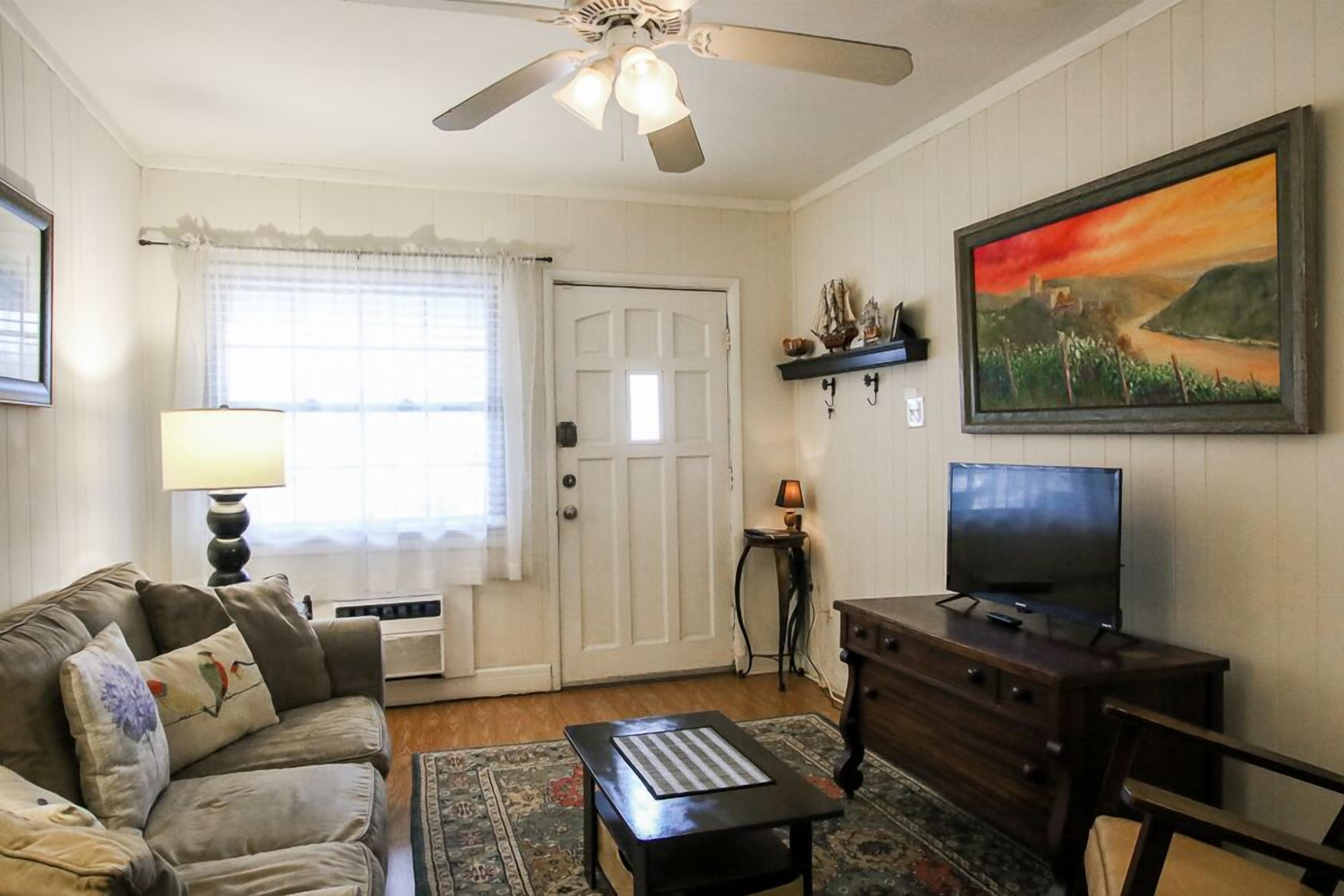A living room with a sofa, a coffee table, a TV on a wooden stand, a ceiling fan, a door, and a painting on the wall.