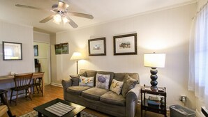 Ein Wohnzimmer mit grauem Sofa, zwei Lampen, gerahmten Bildern an der Wand, einem Deckenventilator und einem kleinen Tisch mit einem Korb.