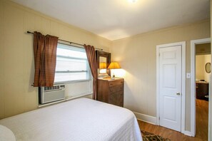 A bedroom with a bed, a dresser, a mirror, a lamp, a window with curtains, and a door. - The Savannah Belle Forsyth Park A Side (Savannah)