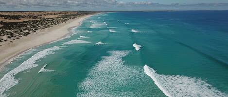 An aerial view of a beach with clear turquoise waters and white waves, sandy shore, and rocky terrain.