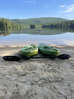Deux kayaks et une pagaie sur une plage de sable, au bord d’un lac paisible, avec un bateau visible au loin et des collines boisées en arrière-plan.