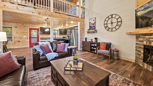 A spacious living area with a high ceiling, wooden flooring, a fireplace, a large clock, and a kitchen in the background.