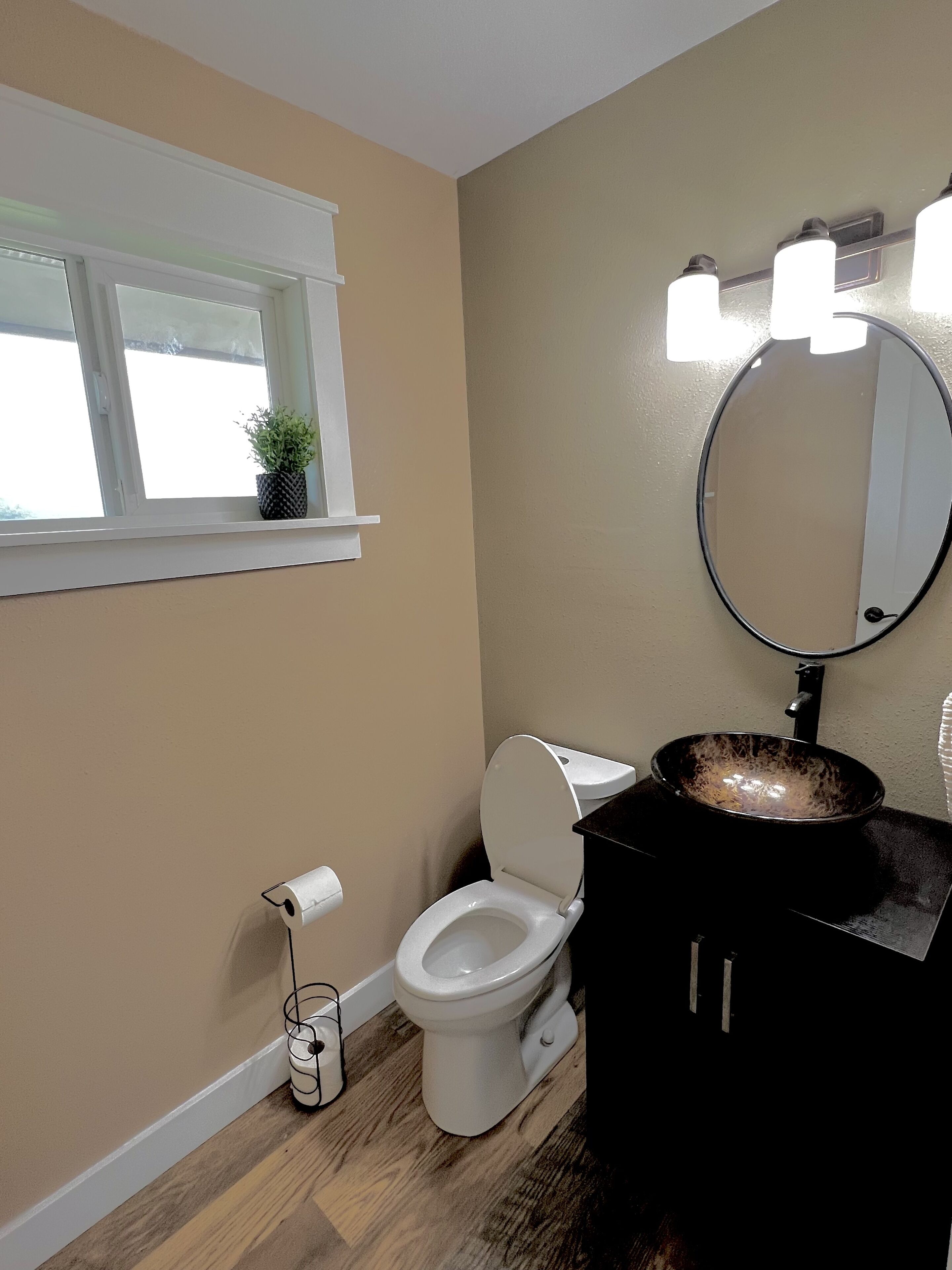 Un baño con inodoro, lavamanos con espejo y una ventana con planta.