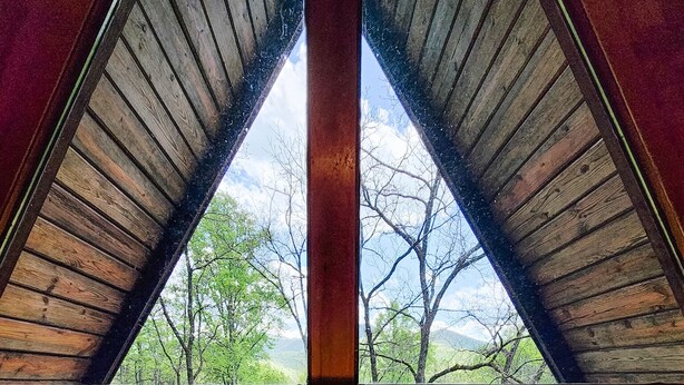 A room with a wooden ceiling and a large window. There are two lamps on a table, and a book titled "World Atlas" is visible. - Dog Friendly Smoky Mts A-Frame with Views and Modern Decor (Sylva)