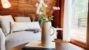 A cozy room with a wooden ceiling, a sofa with cushions, a white vase with flowers, and a view of a wooden deck outside. - Dog Friendly Smoky Mts A-Frame with Views and Modern Decor (Sylva)