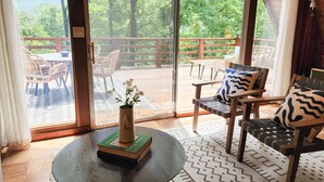 A room with a large sliding glass door leading to an outdoor deck with chairs and a table. A book and a vase with flowers are on a round table in the room. - Dog Friendly Smoky Mts A-Frame with Views and Modern Decor (Sylva)
