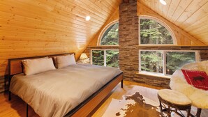 Un dormitorio con techo de madera, pared de piedra, cama grande, dos ventanas y vistas a árboles.