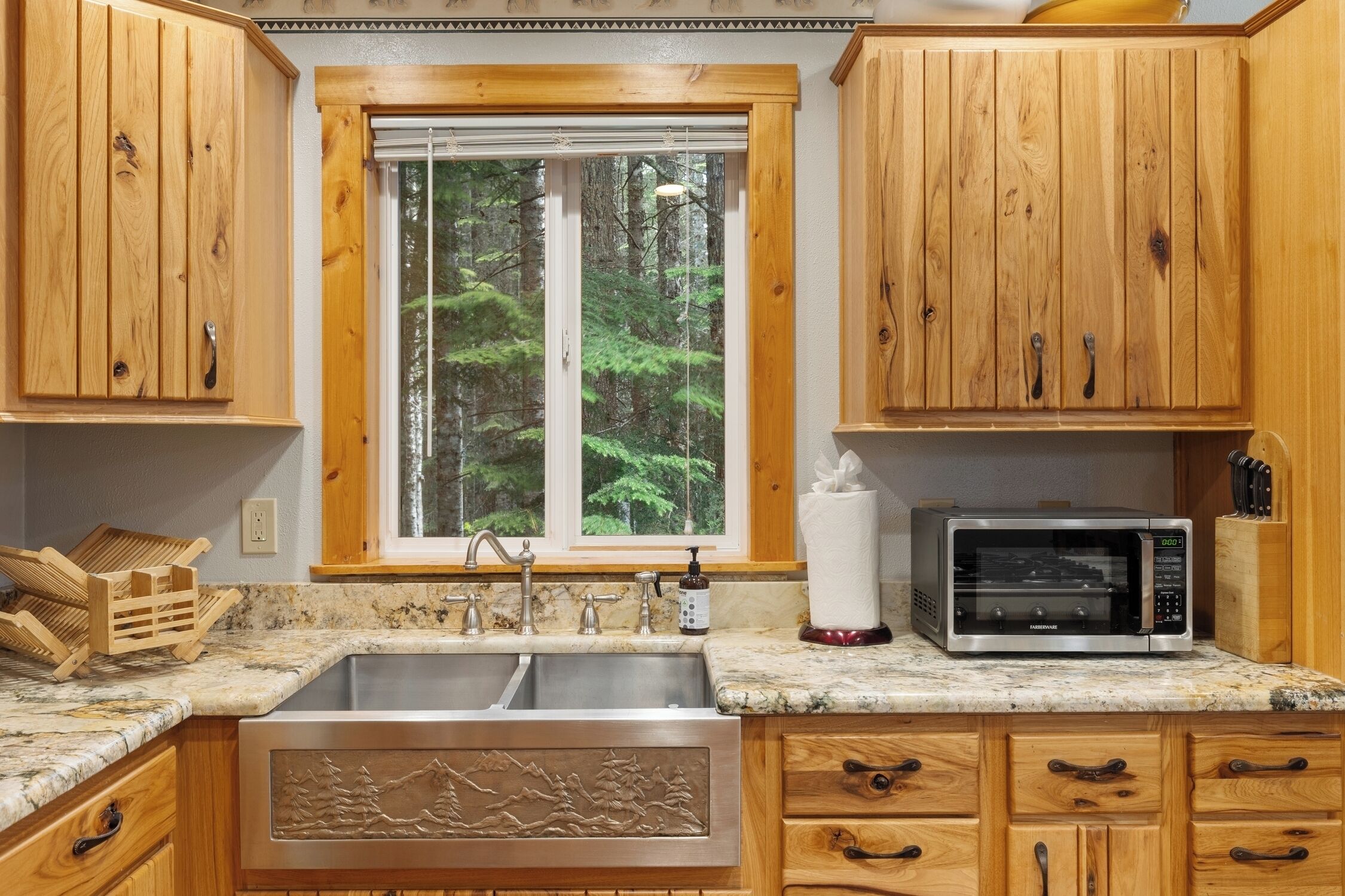 A kitchen with wooden cabinets, a stainless steel sink, a microwave, and a window with a forest view.