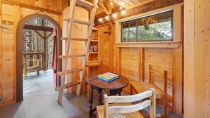 Interior de una cabaña de madera con una mesa redonda, dos sillas, una escalera y un ventanal con vistas a un bosque.
