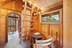 Interior de una cabaña de madera con una mesa redonda, dos sillas, una escalera y un ventanal con vistas a un bosque.