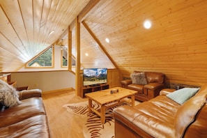 Interior acogedor de una cabaña de madera, con techo de madera, un televisor, sofás de cuero y una mesa de centro de madera.