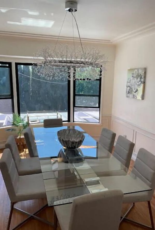 Uma área de refeições com mesa de vidro rodeada por seis cadeiras, um lustre decorativo e grandes janelas com vista para o exterior.