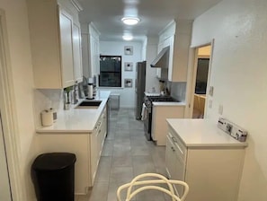 Uma cozinha estreita com armários brancos, um cesto de lixo preto e uma cadeira branca.