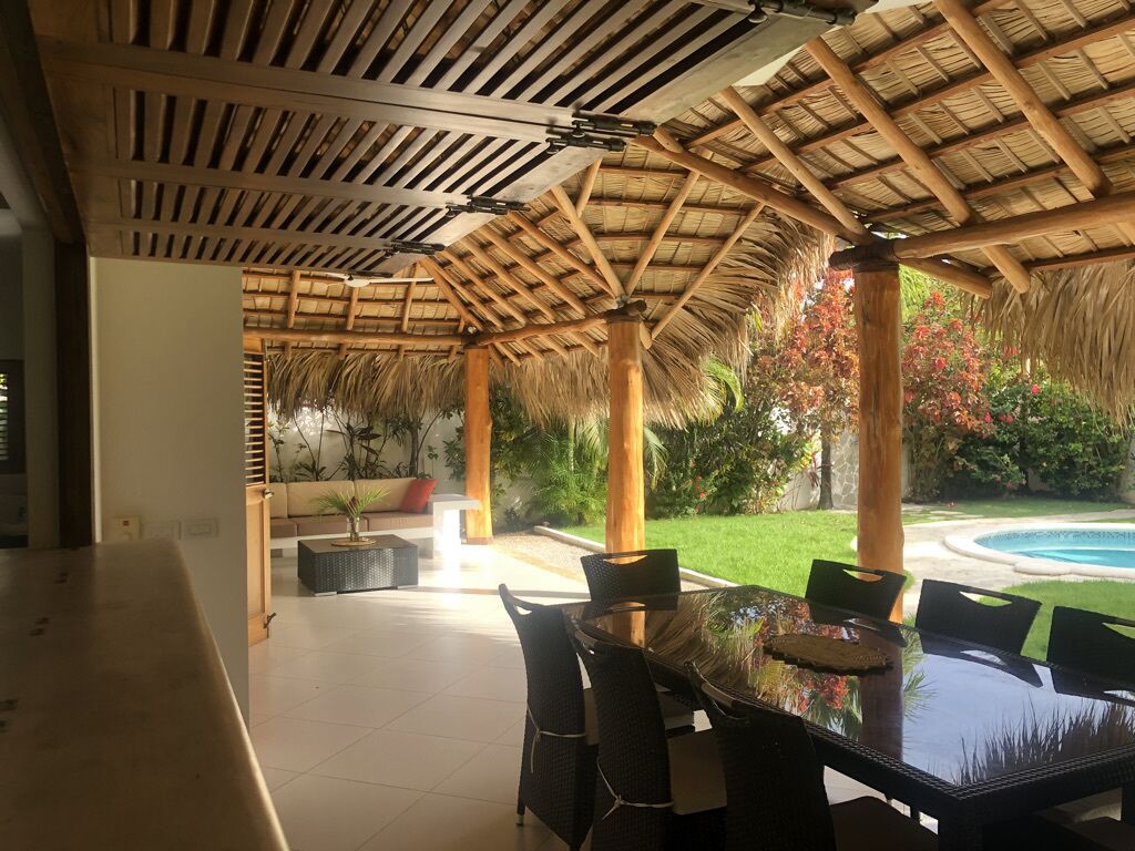 A covered outdoor dining area with a glass table and chairs, surrounded by a wooden pergola and lush greenery.