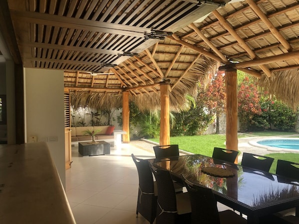A covered outdoor dining area with a glass table and chairs, surrounded by a wooden pergola and lush greenery.