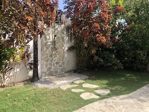 Parede de pedra com chuveiro integrado, rodeada por um jardim com grama e pedras no caminho.