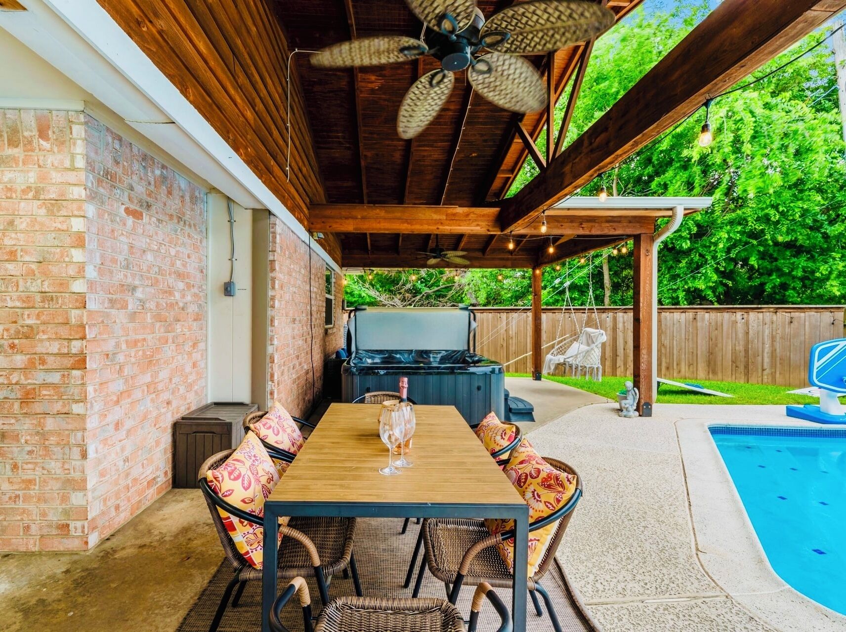 An outdoor patio with a wooden table and wicker chairs, a hot tub, and a swimming pool.