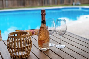A bottle of champagne, two wine glasses, and a wicker basket on a wooden table by a pool. - Villa D’Oro, Minutes to AT&T Stadium, Pool, Spa,Arcade, Game Room (North Richland Hills)