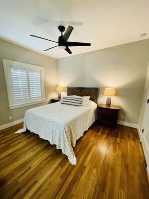 A bedroom with a bed, two nightstands, a ceiling fan, a window with blinds, and wooden flooring.