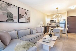A modern living room with a grey sofa, a coffee table, and a fluffy rug. There is a dining area with a table and chairs. The kitchen is visible in the background with modern appliances.