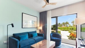A modern hotel room with a blue sofa, a wooden coffee table, a side table, a floor lamp, and a view of a beach and palm trees through large windows.