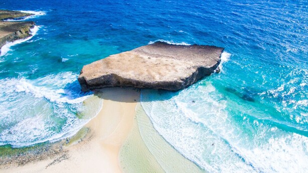 A beach with a large rock formation surrounded by clear blue waters.
