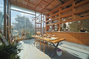 A dining area with a wooden table and chairs, a large window with a view of trees, and wooden shelves with various items.