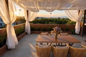An outdoor patio with a wooden table and wicker chairs, draped with white curtains, overlooking a garden and cityscape. - Dali Village Inn (Dali City)