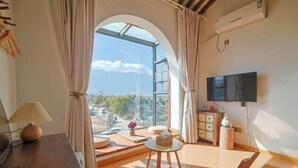 A room with a large arched window, a flat-screen TV, a wooden table, and a sofa. - Dali Village Inn (Dali City)