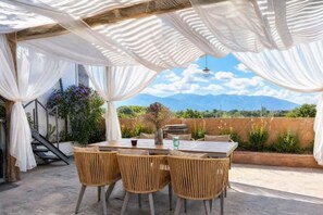 An outdoor dining area with a table and chairs, a pergola with white fabric, and a mountainous landscape in the background. - Dali Village Inn (Dali City)