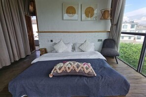 A bedroom with a bed, pillows, a chair, and a view of greenery through large windows. - Dali Village Inn (Dali City)