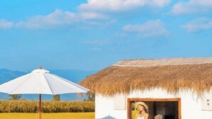 A thatched-roof hut with a white exterior, a white umbrella, and a view of a field and mountains in the background. - Dali Village Inn (Dali City)