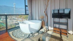 A modern bathroom with a large bathtub, a television on a stand, and a view of the mountains through a large window. - Dali Village Inn (Dali City)