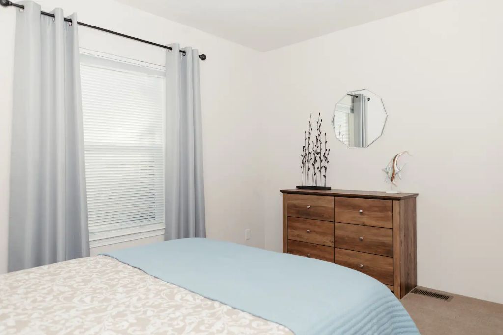 A bedroom with a bed, a dresser, a mirror, and a window with curtains.
