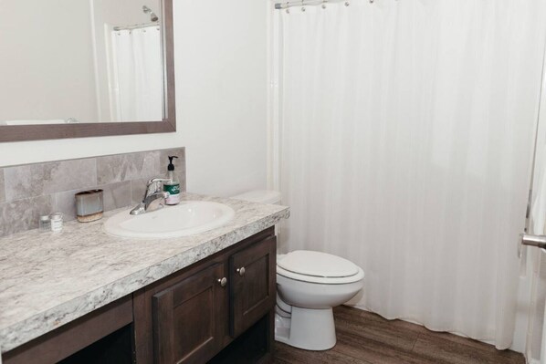 A bathroom with a marble sink, dark wooden cabinets, a white toilet, and a shower curtain. - Neptune Haven Getaway Near the Ocean (Ferndale)