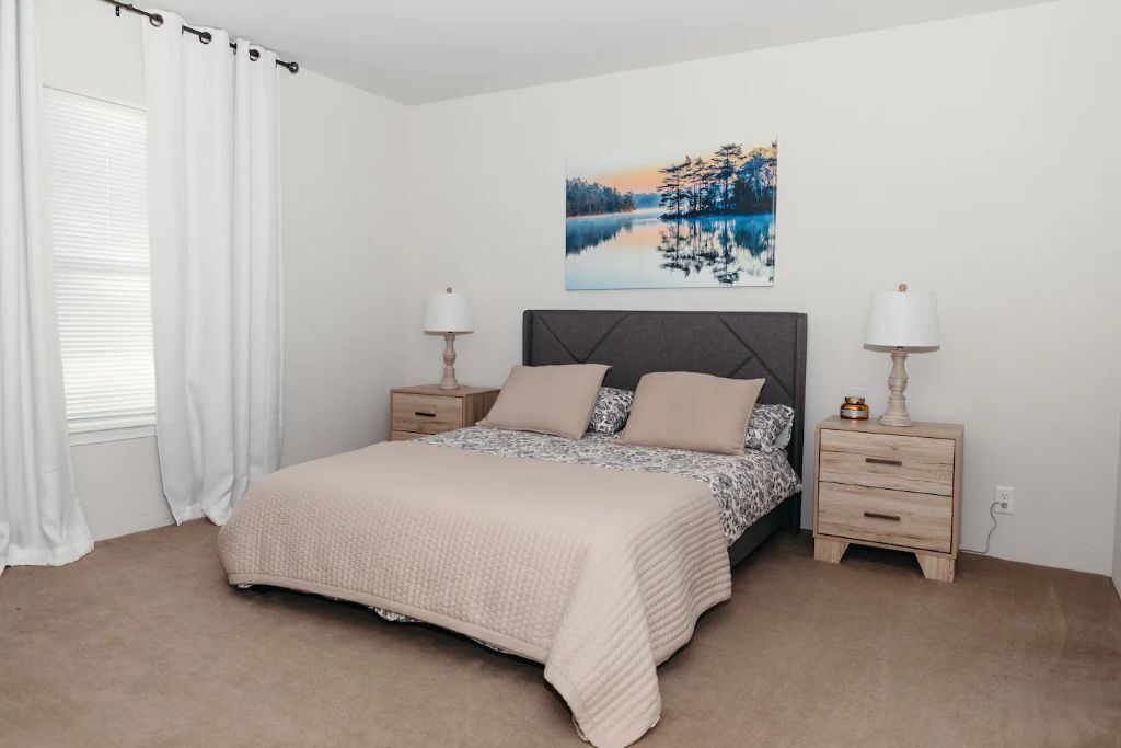 Une chambre à coucher avec un lit, deux tables de chevet, une commode, une fenêtre avec des rideaux et un tableau au mur.