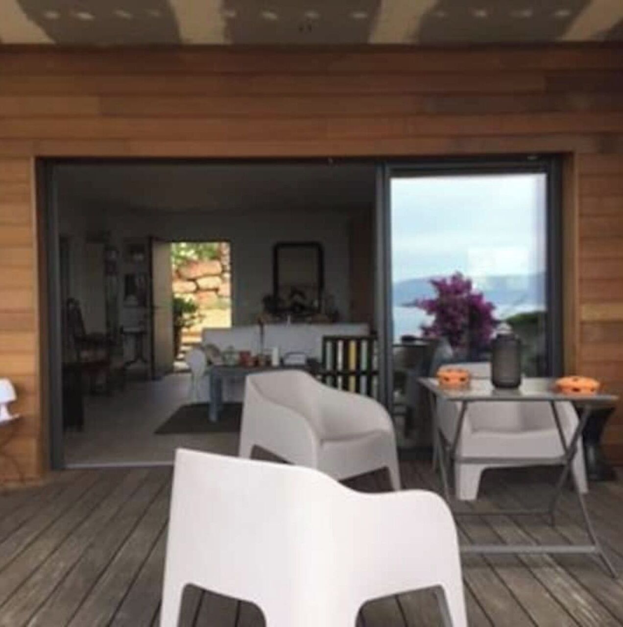 Un patio abierto con cubierta de madera, sillas blancas y una mesa con un jarrón de flores.