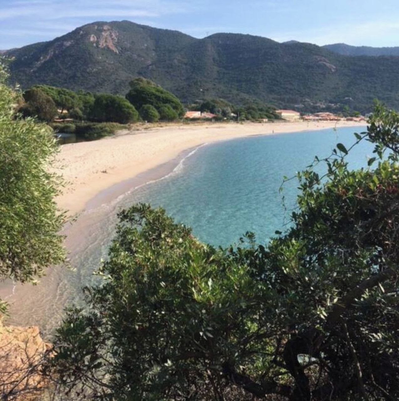 A coastal landscape with a sandy beach, clear blue waters, and green hills in the background.