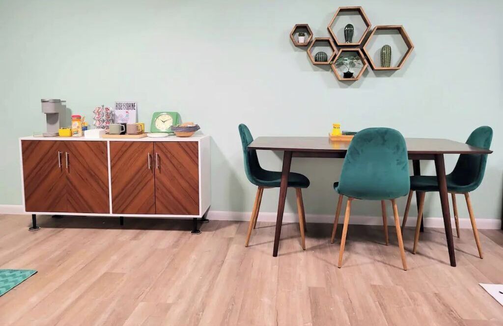 A dining area with a wooden cabinet, teal chairs, and a hexagonal wall decoration.