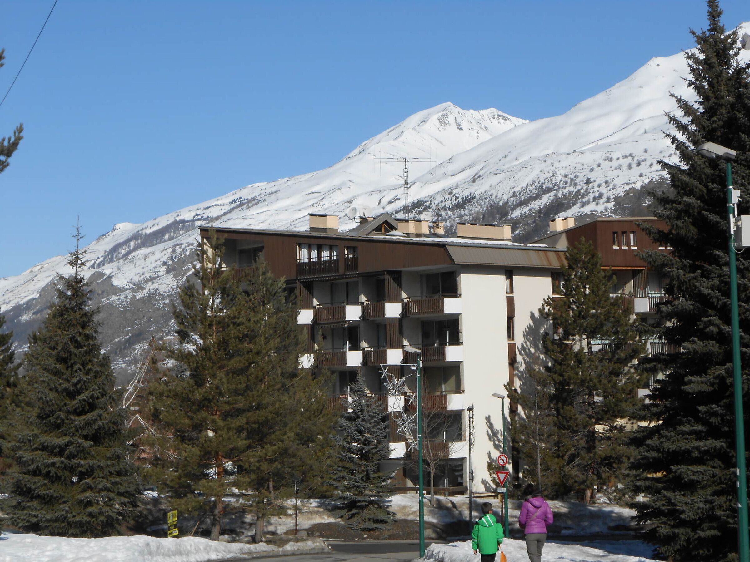 A multi-story building with balconies, surrounded by snow-covered mountains and pine trees.