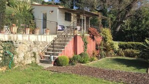 Una casa de dos pisos con una escalera que conduce al nivel superior. Cuenta con un pequeño porche con barandilla y una silla. La casa está rodeada por un jardín con diversas plantas y césped.