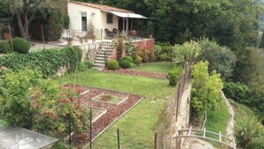 Una casa con porche cubierto, un jardín con césped y parterres de flores, y un muro de piedra.