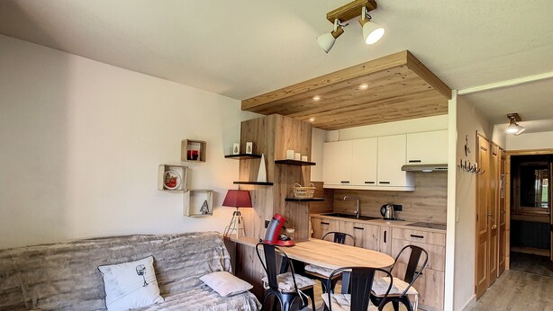 A cozy living room with a sofa, a dining table, and chairs. There is a kitchen area with wooden cabinets and a red lamp. - Charming 2-room apt in Combloux with terrace, parking and ski locker (Combloux)