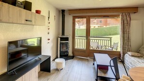 A cozy living room with a TV, a fireplace, a dining table with chairs, and a balcony with a view of the outside. - Charming 2-room apt in Combloux with terrace, parking and ski locker (Combloux)