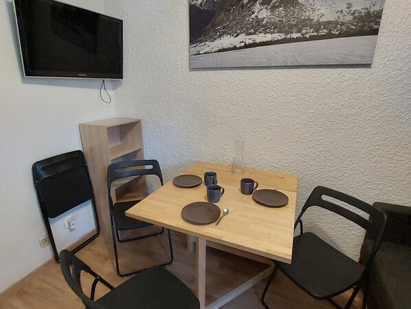 Un petit coin repas avec une table en bois, quatre chaises, un téléviseur fixé au mur et un tableau représentant un paysage enneigé.