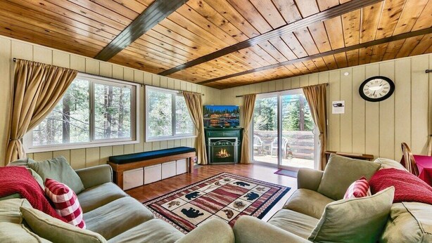 A cozy living room with a wooden ceiling, large windows, a fireplace, and a clock on the wall. - Cozy Forest Getaway | Foosball Fun + A/C Comfort (South Lake Tahoe)